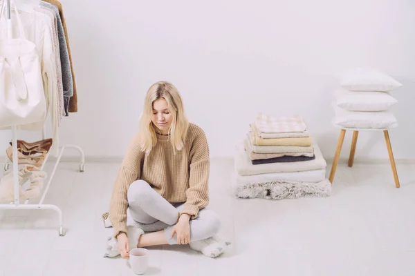 Jonge vrouw zit op de vloer te genieten van koffie en ochtend routine in de buurt van stapel handdoeken kussens, plaid. Huis in Scandinavische stijl, kledingrek in moderne lege garderobe. — Stockfoto