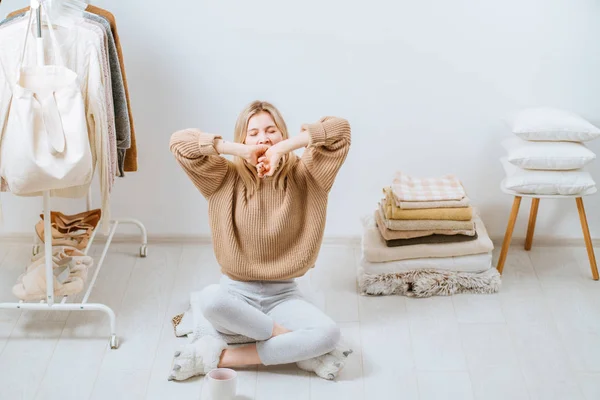 Young woman sitting on the floor near stack of towels pillows, cover plaid on white wall stack tower next to her on chair. Home in scandinavian style, clothes rack in modern empty wardrobe room. — 스톡 사진