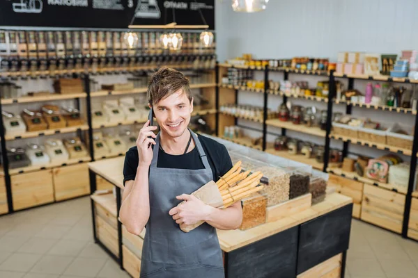 Professional caucasian bakery owner in grey apron holding grissini and smiling inviting guests to own bakery shop.Positive male owner standing over grocery shop showing hospitality. Small business.