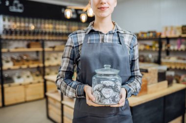 Sıfır Atık Dükkanında Karışık Yarış Kadını Süper Gıda Satıyor. Markette ayakta duran cam şişelerde bir sürü sağlıklı yiyecek var. Plastik Bilinçli Minimalizm Vegan Yaşam Tarzı Konsepti yok. Seçici odak