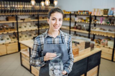 Sıfır Atık Dükkanında Karışık Yarış Kadını Süper Gıda Satıyor. Markette ayakta duran cam şişelerde bir sürü sağlıklı yiyecek var. Plastik Bilinçli Minimalizm Vegan Yaşam Tarzı Konsepti yok. Seçici odak
