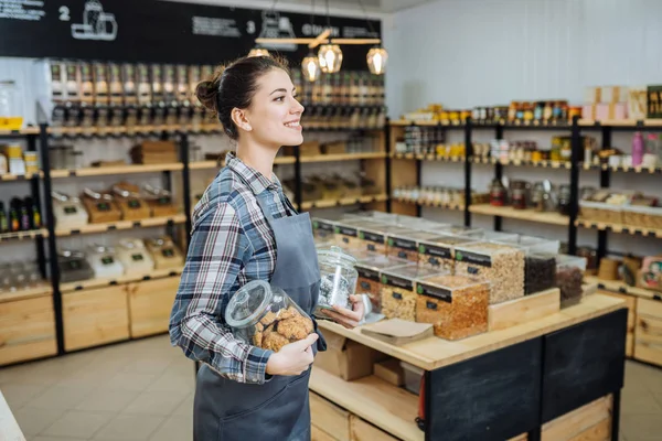 Proprietario di razza mista che vende superprodotti nel negozio Zero Waste Shop. Un sacco di cibo sano in bottiglie di vetro in stand nel negozio di alimentari. Nessun minimalismo cosciente di plastica Vegan Lifestyle Concept. Focus selettivo — Foto Stock