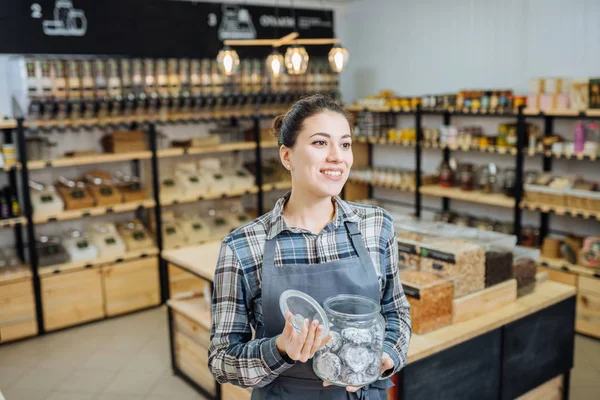 Proprietario di razza mista che vende superprodotti nel negozio Zero Waste Shop. Un sacco di cibo sano in bottiglie di vetro in stand nel negozio di alimentari. Nessun minimalismo cosciente di plastica Vegan Lifestyle Concept. Focus selettivo — Foto Stock