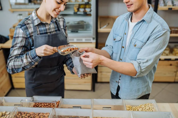 Giovane donna sorridente con un sacchetto di carta in mano sceglie le nocciole e le versa in un sacchetto . — Foto Stock