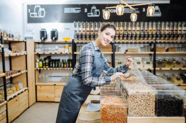 Sıfır Atık Dükkanında Karışık Yarış Kadını Süper Gıda Satıyor. Markette ayakta duran cam şişelerde bir sürü sağlıklı yiyecek var. Plastik Bilinçli Minimalizm Vegan Yaşam Tarzı Konsepti yok. Seçici odak