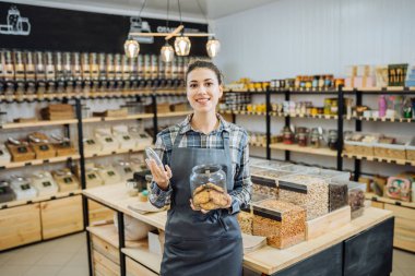 Sıfır Atık Dükkanında Karışık Yarış Kadını Süper Gıda Satıyor. Markette ayakta duran cam şişelerde bir sürü sağlıklı yiyecek var. Plastik Bilinçli Minimalizm Vegan Yaşam Tarzı Konsepti yok. Seçici odak