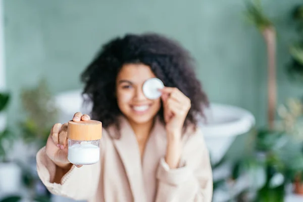 Positief vriendelijke multi-etnische zwarte krullende vrouw in beige badjas, bedekking oog, met behulp van katoen pad presenteren crème of lotion product. Geen afval concept. Groene en bewuste levensstijl concept — Stockfoto