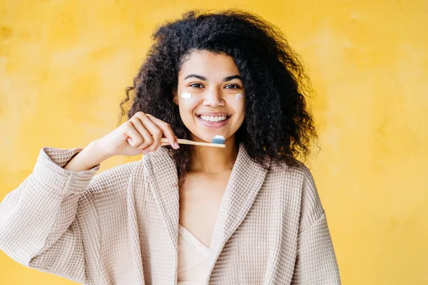 Portrait of African American woman dressing gown brushes teeth on yellow background. Dental hygiene, morning routine concept. Plastic less, zero waste concept, mixed race multiethnic woman