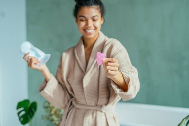 Neşeli, enerjik genç bir kadın kritik günlerde regl bardağı kullanmayı öneriyor. Bir elinde yeşil banyo arka planında hijyen pedi tutuyor. Çevre dostu, sıfır atık, konsepti yeniden kullan.