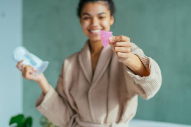 Neşeli, enerjik genç bir kadın kritik günlerde regl bardağı kullanmayı öneriyor. Bir elinde yeşil banyo arka planında hijyen pedi tutuyor. Çevre dostu, sıfır atık, konsepti yeniden kullan.