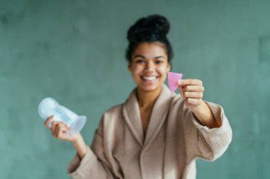 Neşeli, enerjik genç bir kadın kritik günlerde regl bardağı kullanmayı öneriyor. Bir elinde yeşil banyo arka planında hijyen pedi tutuyor. Çevre dostu, sıfır atık, konsepti yeniden kullan.
