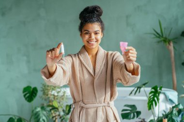 Çevre dostu Afro-Amerikalı kadın iki elinde tampon ve regl bardağı tutuyor, yan bakıyor, banyo içinde bej bornoz giyiyor, karşılaştırıyor. Regl, sıfır atık, yeniden kullanım kavramı.
