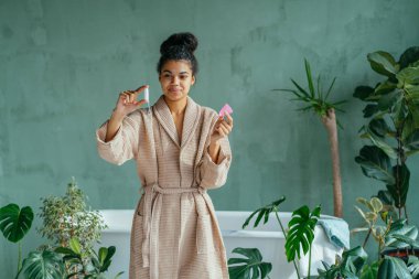 Pozitif görünümlü, siyah saçlı, kıvırcık saçlı, çok ırklı, tampon ve adet bardağı tutan, arka plandaki yeşil, samimi tuvalette tampon kullanan bir kadının portresi..