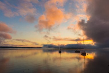 Gölde teknelerle gün doğumu, suda güneşin yansıması, yaz sabahında sis ve bulutlar.
