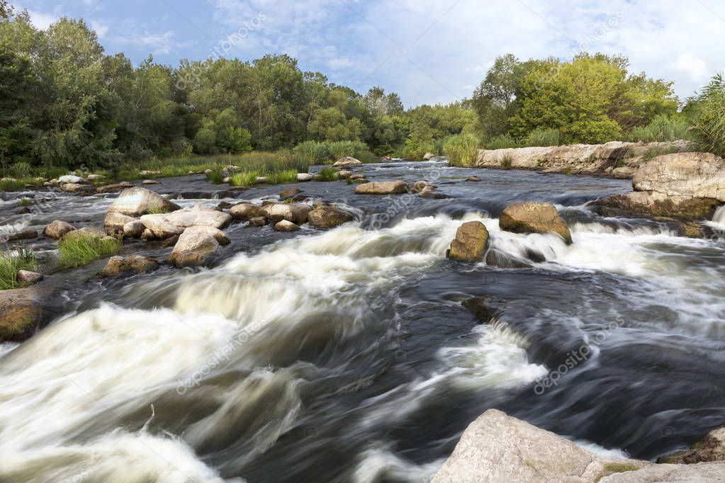 The rapid flow of the river, rocky coasts, rapids, bright green vegetation and a cloudy blue summer sky