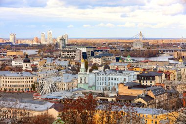The landscape of the autumn city of Kyiv overlooking the old district of Podil with a Ferris wheel and a bell tower with a gilded dome, the Dnipro River and many bridges.