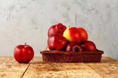 Olgun kırmızı elmalar, gri beton arka planı olan eski ahşap bir masada hasır bir sepetin içinde yatıyor..