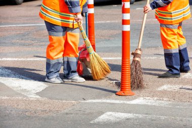 Turuncu üniformalı kadın hademeler caddeyi tarıyor ve yoldaki ve turuncu yol sütunlarındaki çitlerin yanındaki çöpleri temizliyorlar..