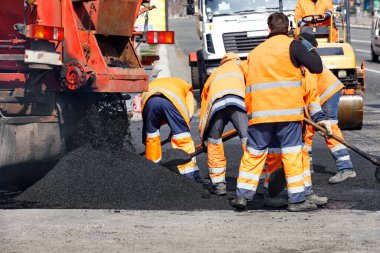 Yol işçilerinden oluşan ekip yolun bir kısmını taze asfalt ile güncelliyor ve yol yüzeyini eşit olarak dağıtmak için küreklerle düzleştiriyor..