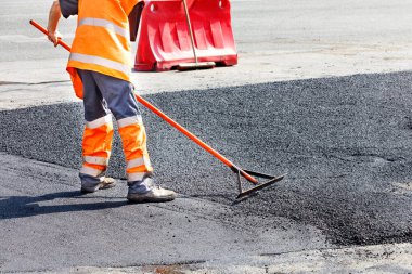 A road service worker updates a stretch of road with fresh hot asphalt and smoothes it with a metal level for manual repairs. clipart