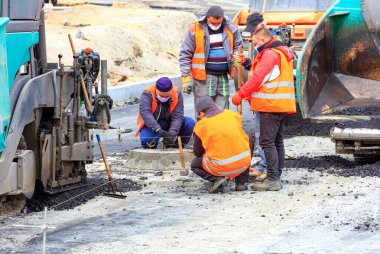 At a new work site, among large pavers, a workers team in protective masks discusses and sets the necessary level for asphalting the road. clipart