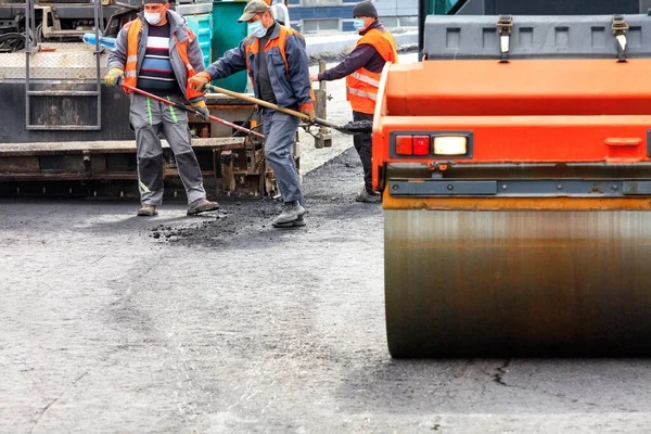 Koruyucu tıbbi maskeler ve tulumlarla çalışan yol işçilerinden oluşan bir ekip, güçlü yol ekipmanları ve kürekler kullanarak çalışma alanına yeni bir asfalt kaplama döşedi..