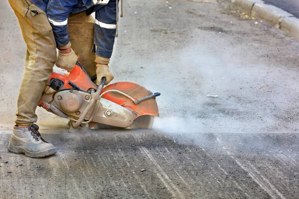Taşınabilir beton kesici kullanan ve elmas bıçak kesen bir işçi yolun bir kısmını onarmak için toz bulutunda yıpranmış asfalt kesiyor..