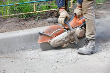 Atık benzin testeresi kullanan bir yol işçisi beton toz bulutu ve uçan kıvılcımlarla asfaltı kesiyor..