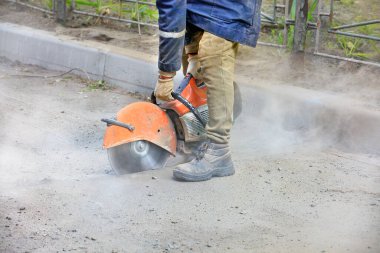 Taşınabilir benzin kesici kullanan ve elmas bıçak kesen bir işçi eski asfaltı toz bulutu ve uçan kıvılcımlarla keser..
