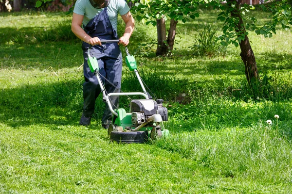 庭師は緑の芝生の世話をし 春の庭の木の間に工業用ガソリン粉で緑の草を刈る — ストック写真