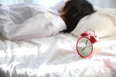 Asyalı tembel kadın beyaz yatak odasındaki saatle yatakta uyanıyor.
