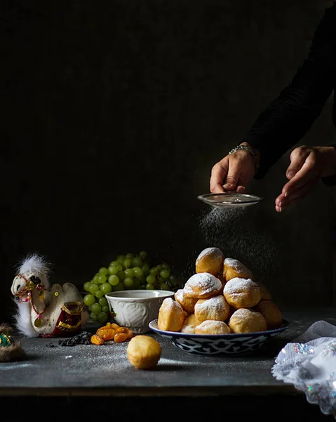 Kazakh baursaks on a plate with patterns. Women\'s hands sprinkle them with powdered sugar. On the table are grapes, dried fruits, and a camel.