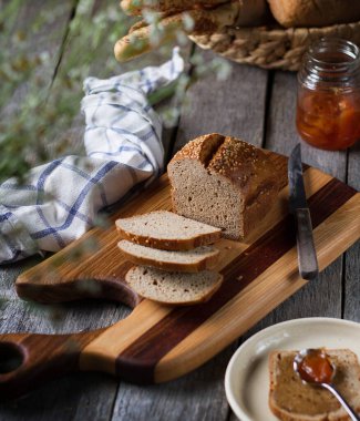 Tahtada dilimlenmiş ekmek eski bir masada, bir kavanoz kayısı reçeli, bir havlu ve çiçekler.