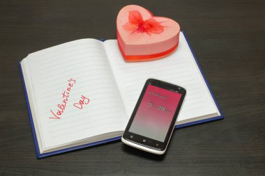 14 Şubat Sevgililer Günü 'nde bir not defterine cep telefonu ve siyah tahta bir masanın üzerinde bir kurdeleyle kalp şeklinde bir kutu yazmak.