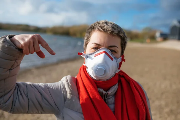 Portrait of Woman wearing white and red a face mask and red scarf, pointing at your mask outdoors