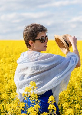 Güneş gözlüğü takan ve mavi atkı takan bir kadın güneşli bir bahar gününde sarı kolza tohumu tarlasında şapkasını çıkarıyor.