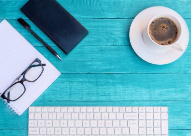 Beyaz kahve, beyaz klavye, siyah defter, siyah kalem, siyah bardaklar ve turkuaz ahşap masadaki beyaz boş çarşaf. İnsansız Düz Yat. Yukarıdan görüntüle.
