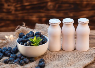 Beyaz kâsesinde yaprak olan taze yaban mersini, kahverengi ahşap arka planda üç yoğurt şişesi.