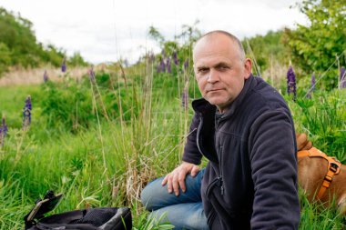  Siyah ceketli ve kot pantolonlu bir adam yeşil çayırda oturuyor ve telefonda fotoğraf çekiyor.