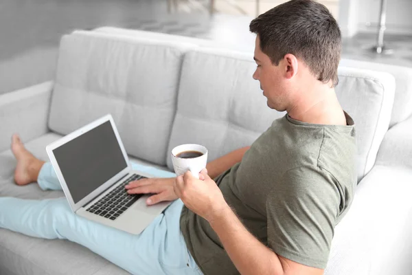 Hombre guapo sentado en el sofá con portátil y taza de café —  Fotos de Stock