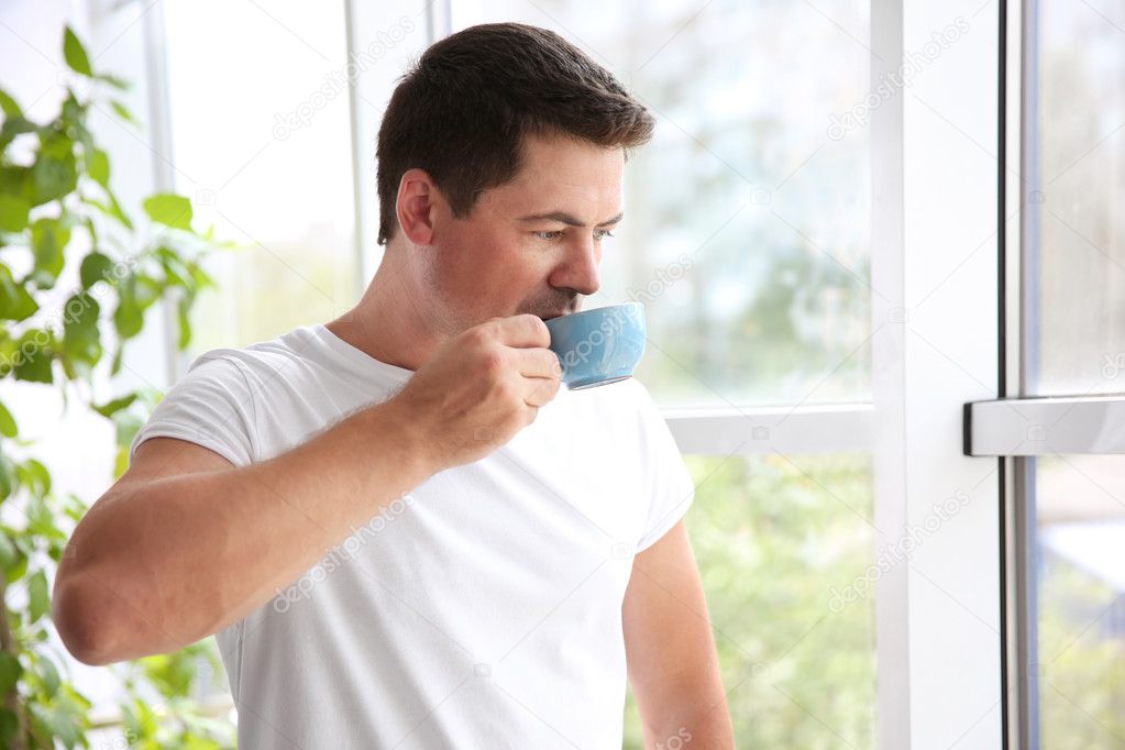 Handsome man standing near window with cup of morning coffee