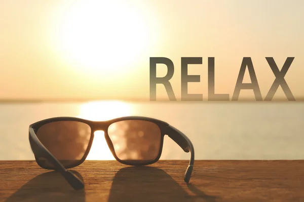 Sunglasses on wooden table and word RELAX on blurred sunset background. — Stock fotografie