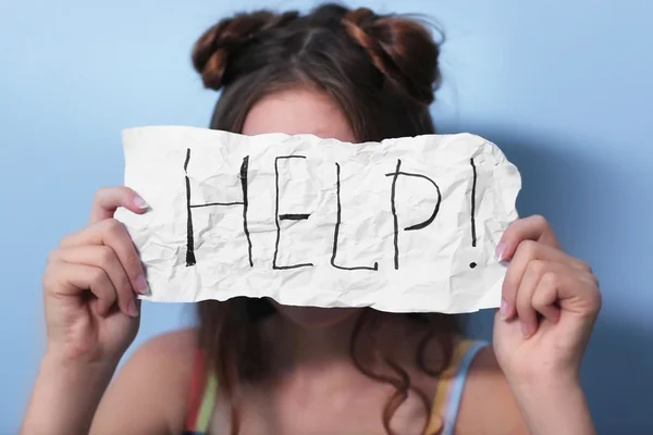 Teenager girl holding paper with HELP text on blue wall background