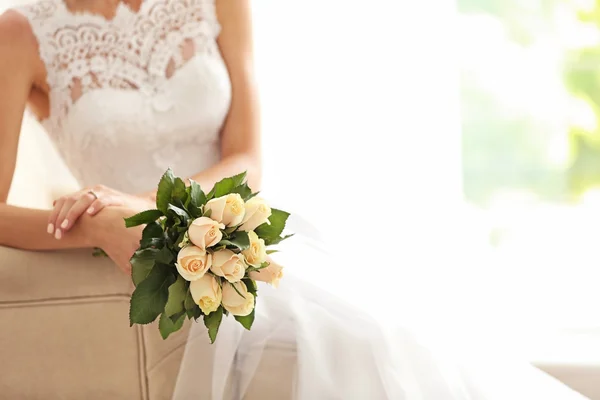 Bride in beautiful dress with wedding bouquet sitting on sofa — Stock Photo, Image