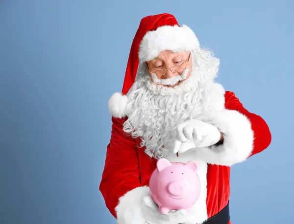 Santa Claus poniendo moneda en alcancía sobre fondo azul — Foto de Stock