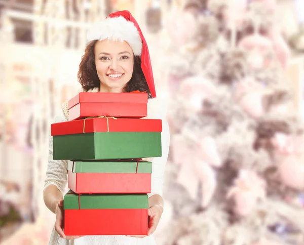 Mulher feliz bonita em chapéu de Santa com presentes de Natal em fundo de abeto decorado borrado. Conceito de compras de Natal . — Fotografia de Stock