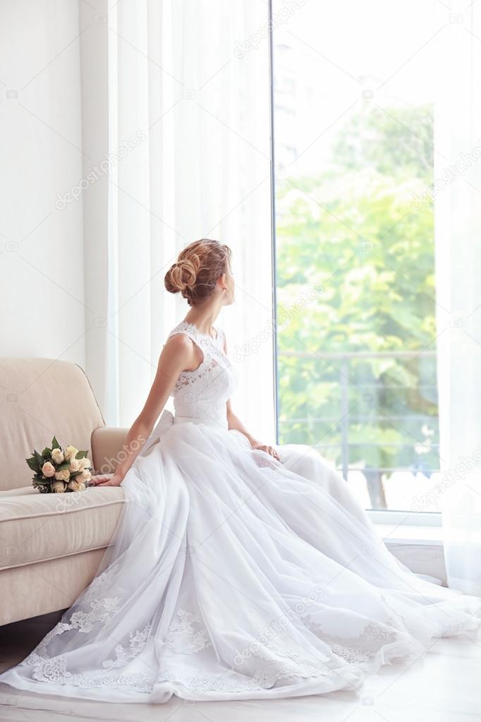 Bride in beautiful dress with wedding bouquet sitting on sofa