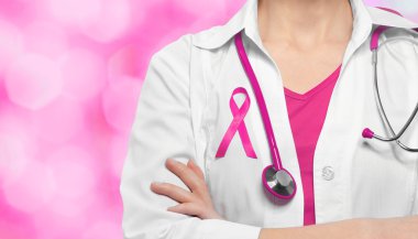 Female doctor with pink ribbon on chest against color background. Oncology and breast cancer concept. clipart
