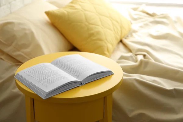 Livro aberto na mesa de cabeceira amarela perto da cama amassada — Fotografia de Stock