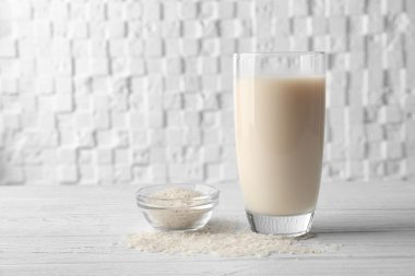 Glass of rice milk and bowl with grains on wooden table against white blurred background clipart